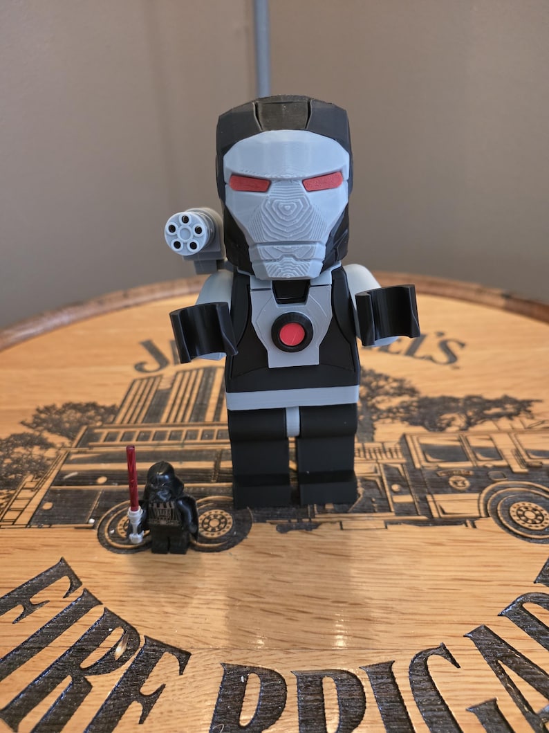 Peut inclure: Une figurine grise et noire de War Machine avec des d&eacute;tails rouges sur les yeux et un canon mont&eacute; sur l'&eacute;paule. Une petite figurine de Dark Vador avec un sabre laser rouge est &eacute;galement pr&eacute;sente. Les figurines sont sur une surface en bois.