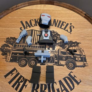 Peut inclure: Une figurine articul&eacute;e grise et noire d&eacute;mont&eacute;e, comprenant un casque, un torse, des bras, des jambes et un pistolet, est dispos&eacute;e sur une surface en bois. La surface pr&eacute;sente le texte "Jack Daniel's" et un dessin de camion de pompiers, avec le texte "Fire Brigade".