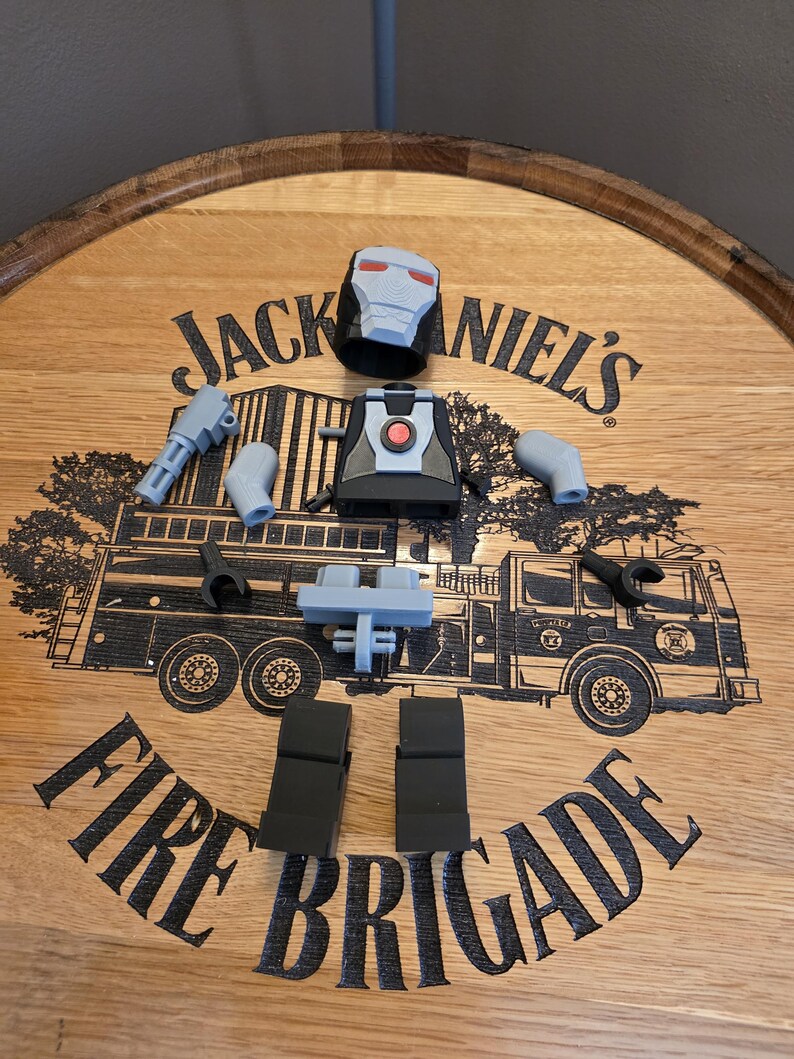 Peut inclure: Une figurine articul&eacute;e grise et noire d&eacute;mont&eacute;e, comprenant un casque, un torse, des bras, des jambes et un pistolet, est dispos&eacute;e sur une surface en bois. La surface pr&eacute;sente le texte "Jack Daniel's" et un dessin de camion de pompiers, avec le texte "Fire Brigade".