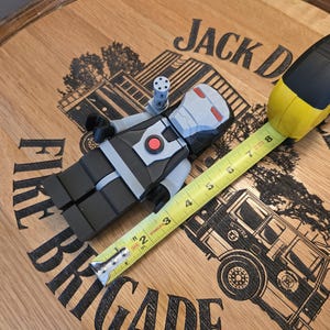 Peut inclure: Une figurine grise, noire et rouge ressemblant &agrave; War Machine, avec une petite fixation de pistolet, est pos&eacute;e &agrave; c&ocirc;t&eacute; d'un m&egrave;tre ruban jaune et noir. La figurine est sur une surface en bois avec les mots "Jack Daniel's" et "Fire Brigade" visibles.