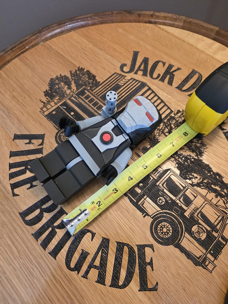 Peut inclure: Une figurine grise, noire et rouge ressemblant &agrave; War Machine, avec une petite fixation de pistolet, est pos&eacute;e &agrave; c&ocirc;t&eacute; d'un m&egrave;tre ruban jaune et noir. La figurine est sur une surface en bois avec les mots "Jack Daniel's" et "Fire Brigade" visibles.
