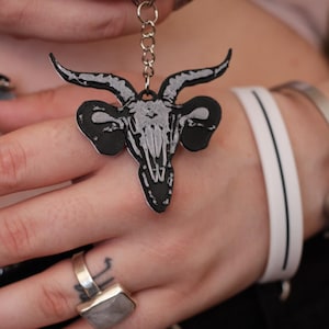 Può includere: Portachiavi con teschio di capra nero e grigio. Il portachiavi presenta un design dettagliato del teschio con corna prominenti, attaccato a un portachiavi color argento. Il teschio è posto su uno sfondo nero, creando un contrasto sorprendente. Accessorio a tema gotico.