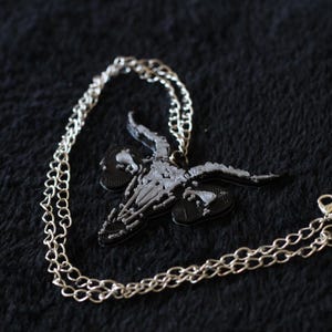 Può includere: Una collana a catena in metallo color argento con un ciondolo a forma di teschio di capra. Il ciondolo è prevalentemente nero con dettagli color argento sulle corna e sulla struttura facciale, appeso a una catena a maglie con moschettone.