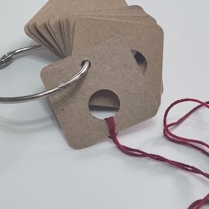 May include: A stack of square, light brown cardboard tags with rounded corners, each featuring a circular hole. The tags are held together by a silver metal binder ring. A single piece of dark red thread is looped through the hole of the front tag.
