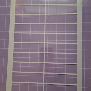 May include: An off-white label sheet with pre-cut rectangular labels, on a purple cutting mat with a grid pattern. The labels are arranged in a grid of 3 columns and 6 rows. The sheet is rectangular.