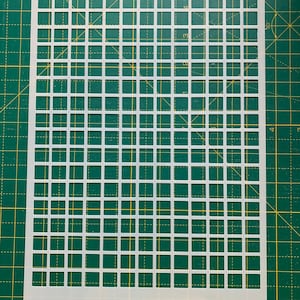 May include: A white plastic stencil with a grid pattern of evenly spaced squares. The stencil is rectangular and has a series of cut-out squares, creating a repeating geometric design. The stencil is placed on a green cutting mat.
