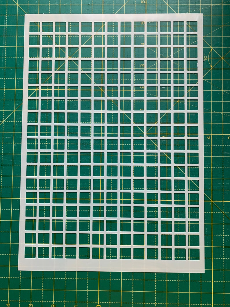 May include: A white plastic stencil with a grid pattern of evenly spaced squares. The stencil is rectangular and has a series of cut-out squares, creating a repeating geometric design. The stencil is placed on a green cutting mat.