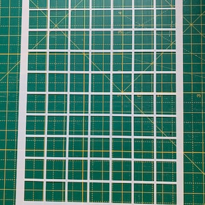 May include: A white plastic stencil with a grid pattern of evenly spaced squares. The stencil is on a green cutting mat with yellow grid lines. The squares are uniform in size and shape, creating a repeating pattern.