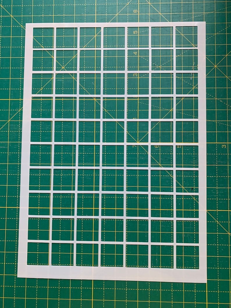 May include: A white plastic stencil with a grid pattern of evenly spaced squares. The stencil is on a green cutting mat with yellow grid lines. The squares are uniform in size and shape, creating a repeating pattern.