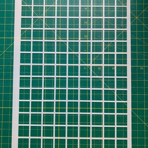 May include: A white plastic stencil with a grid pattern of evenly spaced squares. The stencil is rectangular and lies on a green cutting mat with a grid pattern. The squares are uniform in size and shape, creating a precise grid.