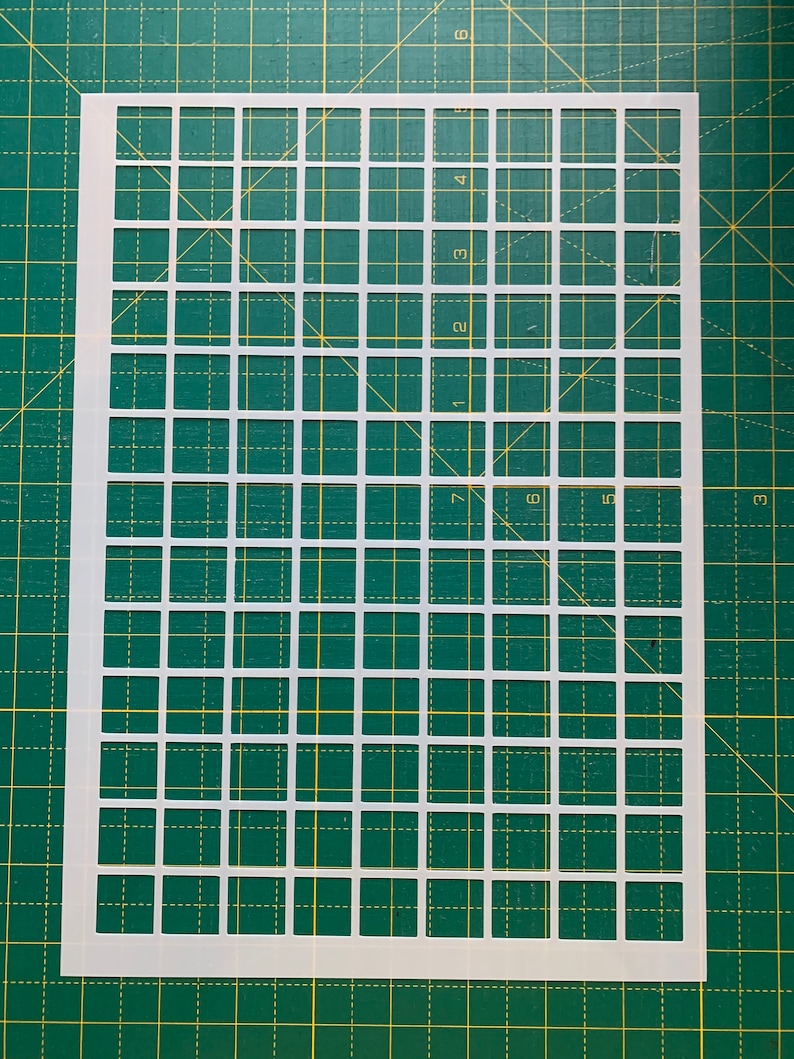 May include: A white plastic stencil with a grid pattern of evenly spaced squares. The stencil is rectangular and lies on a green cutting mat with a grid pattern. The squares are uniform in size and shape, creating a precise grid.