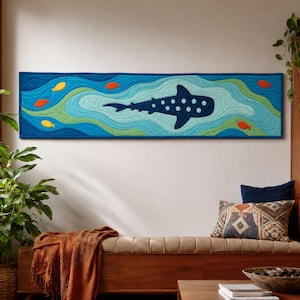 Puede incluir: Un tapiz rectangular largo con un diseño de tela acolchada que muestra un tiburón ballena azul oscuro nadando a través de capas onduladas y estilizadas de agua azul claro, turquesa y verde, acentuado por pequeñas formas de peces naranjas y amarillos.