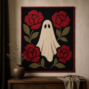 May include: A square wall quilt featuring a central off-white ghost figure with two black oval eyes, surrounded by four large red roses with olive green leaves on a textured black background, all framed by a thin red border.