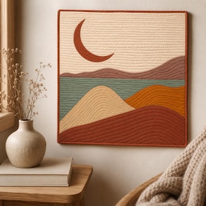 May include: A square quilted wall hanging featuring a landscape design with a crescent moon in the sky above rolling hills. The textile art uses a palette of cream, terracotta, sage green, and mustard yellow with visible horizontal stitching.