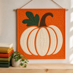 May include: A square quilted wall hanging featuring a large white pumpkin with a dark green leaf and a brown stem, set against a textured orange background. The piece is suspended from a wooden rod with a string, showcasing detailed stitching.