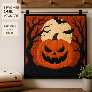 May include: A square fabric wall hanging featuring a quilted orange jack-o'-lantern with a black face, set against a large cream moon and black silhouettes of bare trees and flying bats. The piece has a black border and a bound finish.