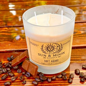 May include: A frosted glass candle with two wicks, labeled "Sun & Moon Scents - Soft Awake." The candle is surrounded by coffee beans and chocolate pieces on a wooden surface. The label mentions coffee, milk, and chocolate.