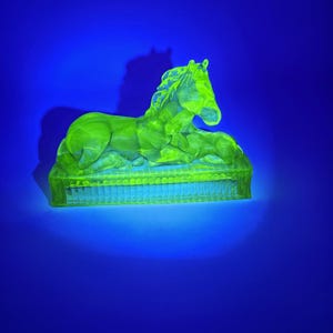Può includere: Una statuetta in vetro verde lime fluorescente raffigurante un cavallo sdraiato su una base rettangolare con bordo scanalato. L'oggetto brilla intensamente sotto la luce ultravioletta, evidenziando il materiale traslucido e la forma scolpita.