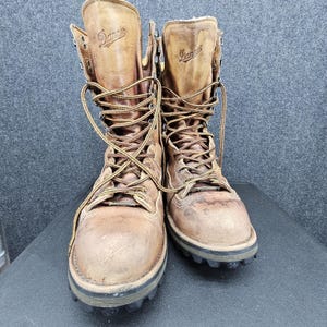 May include: A pair of brown leather work boots with the brand name "Danner" visible on the upper shaft. The boots have a lace-up design with brown and yellow laces, and a thick black sole. The boots show signs of wear.