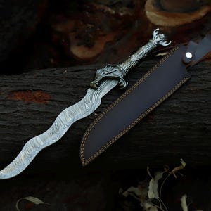 May include: A wavy-bladed dagger featuring a patterned Damascus steel blade and a metallic handle shaped like a dragon head. The dagger is accompanied by a dark brown leather sheath with visible stitching and a silver-coloured metal snap closure.