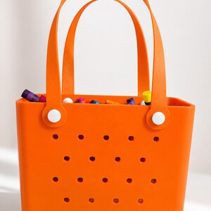 May include: Bright orange tote bag with two arched handles and a series of small holes on the front. The bag is filled with colorful items. White circular fasteners secure the handles to the bag.