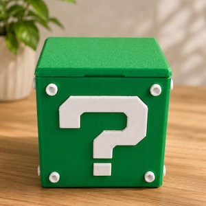 May include: A green box with a white question mark on the front. The box has a textured surface and white circular accents on each corner. The box is sitting on a wooden surface.
