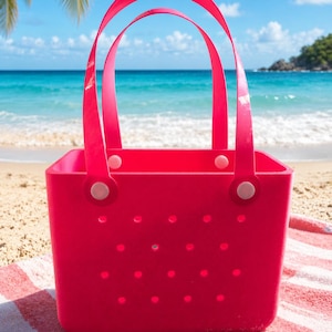 May include: A bright pink tote bag with two arched handles, set on a beach towel with a red and white stripe pattern. The bag has a square shape with small circular holes. The background shows a beach, ocean, and blue sky.