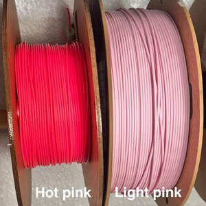 May include: Two spools of 3D printing filament. One spool is hot pink, and the other is light pink. The spools are made of wood and are labeled with the color names. The filament is tightly wound.
