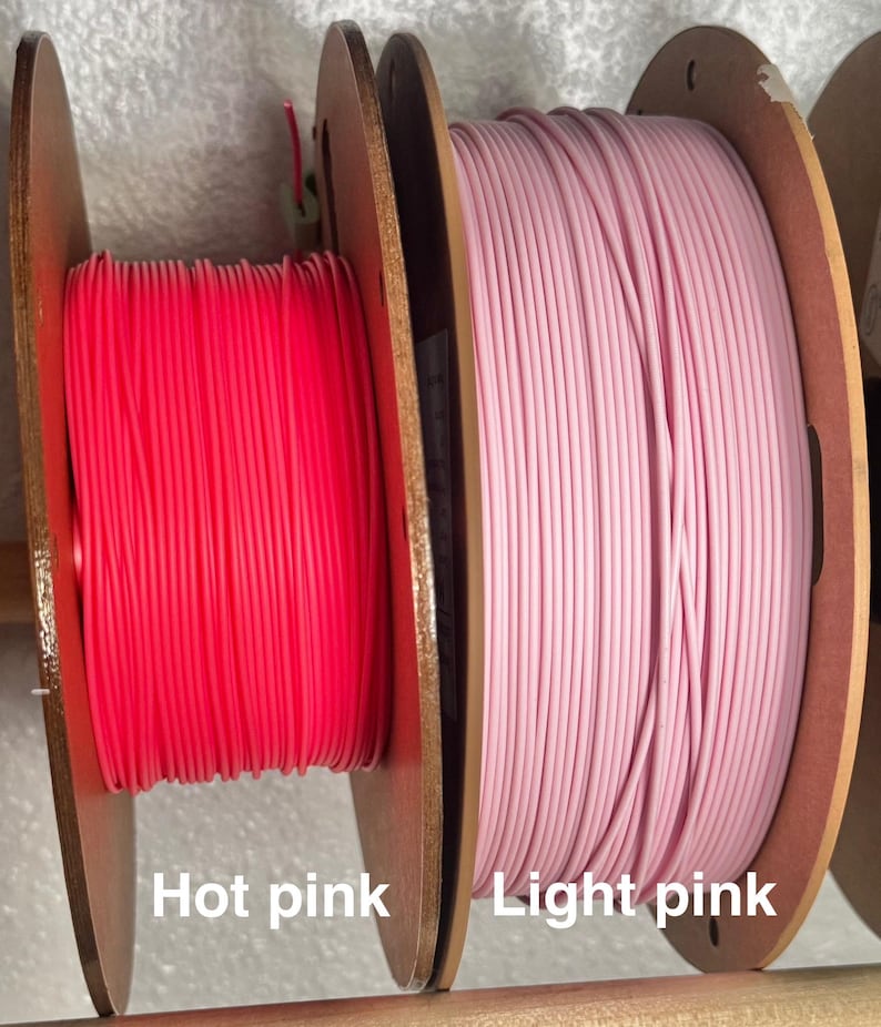 May include: Two spools of 3D printing filament. One spool is hot pink, and the other is light pink. The spools are made of wood and are labeled with the color names. The filament is tightly wound.