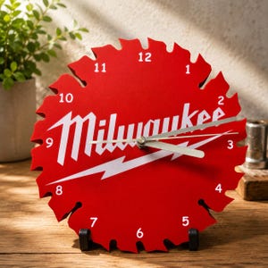 May include: A red clock in the shape of a saw blade, with white numerals and hands. The word "Milwaukee" is displayed in white, with a lightning bolt graphic. The clock is supported by a black stand and sits on a wooden surface.