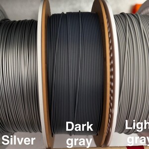 May include: Three spools of 3D printer filament in silver, dark gray, and light gray. The spools are labeled with their respective colors. The filament is tightly wound on wooden spools with white plastic side supports.