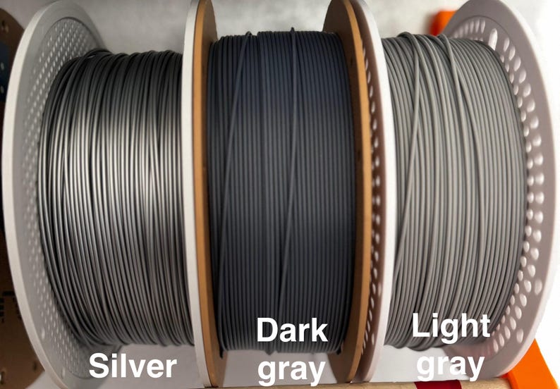 May include: Three spools of 3D printer filament in silver, dark gray, and light gray. The spools are labeled with their respective colors. The filament is tightly wound on wooden spools with white plastic side supports.