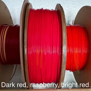 May include: Three spools of red filament are displayed, each wound around a wooden core. The colors range from dark red to bright red, with the text "Dark red, raspberry, bright red" at the bottom.