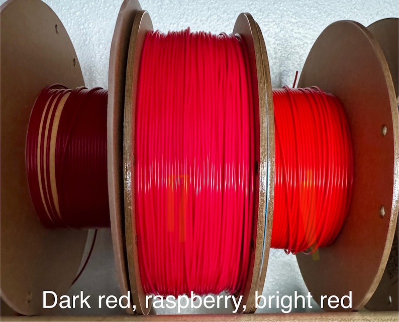May include: Three spools of red filament are displayed, each wound around a wooden core. The colors range from dark red to bright red, with the text "Dark red, raspberry, bright red" at the bottom.