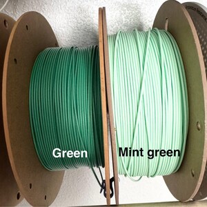 May include: Two spools of wire, one in a deep green and the other in a mint green. The spools are mounted on wooden reels, with the colors labeled "Green" and "Mint green". The wire is tightly wound, suggesting a product for crafting or electrical use.