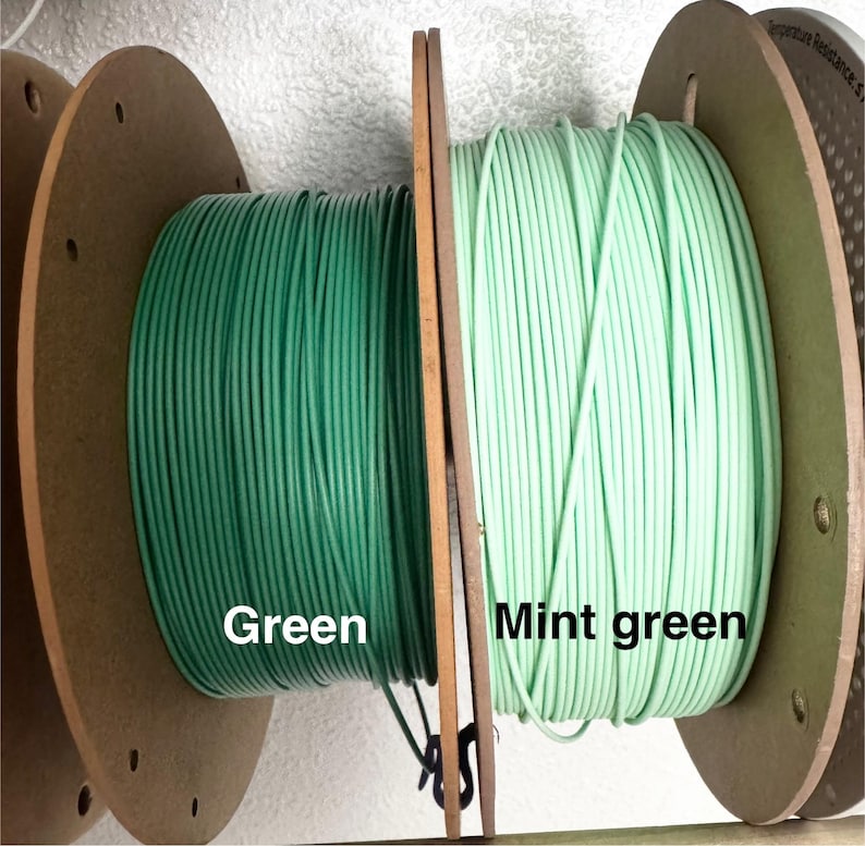 May include: Two spools of wire, one in a deep green and the other in a mint green. The spools are mounted on wooden reels, with the colors labeled "Green" and "Mint green". The wire is tightly wound, suggesting a product for crafting or electrical use.