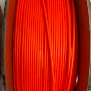 May include: Bright orange filament spool with the word "Orange" printed on the front. The spool is made of a vibrant orange material and is wound around a wooden spool. The image is a close-up shot.