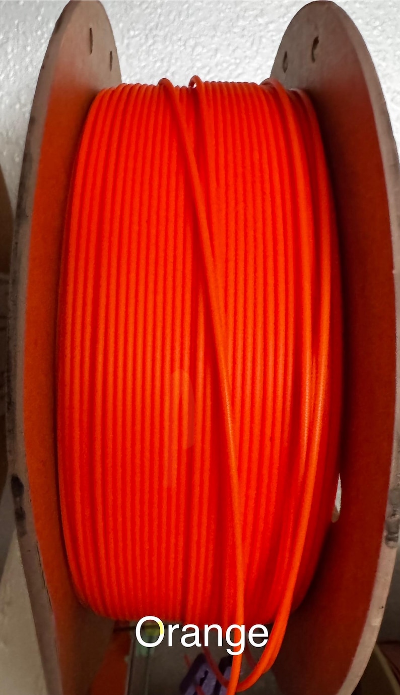 May include: Bright orange filament spool with the word "Orange" printed on the front. The spool is made of a vibrant orange material and is wound around a wooden spool. The image is a close-up shot.