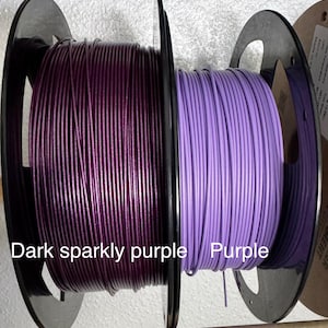 May include: Two spools of filament, one dark sparkly purple and the other purple. The spools are black and the filament is tightly wound. Text on the image reads "Dark sparkly purple" and "Purple".