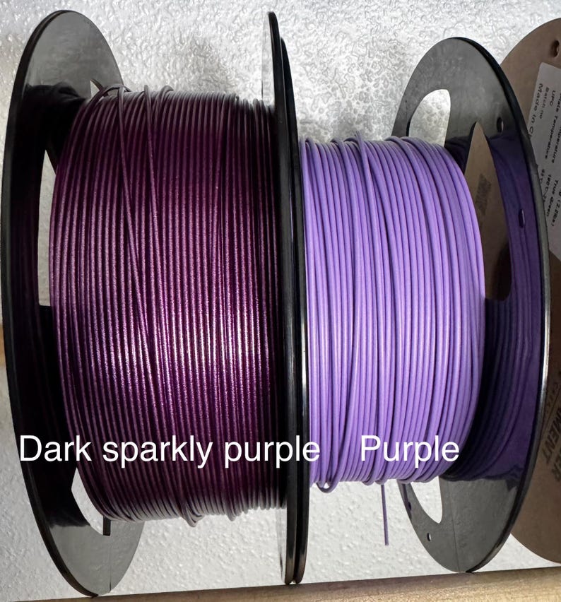 May include: Two spools of filament, one dark sparkly purple and the other purple. The spools are black and the filament is tightly wound. Text on the image reads "Dark sparkly purple" and "Purple".