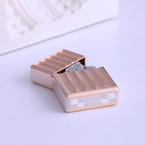 May include: A rose gold metal lighter with a rectangular shape and a hinged lid. The lighter features a textured design on the top and bottom, with a silver Greek key pattern on the sides. The text "SENECA" and "125" are visible.