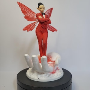 May include: A figurine of a fairy in a red suit with matching wings, standing on a white hand. The fairy has a black bow tie and is posed with arms crossed. The base is a black rotating stand.