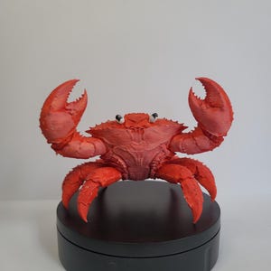 May include: A detailed, red crab sculpture with raised claws and textured shell. The crab is displayed on a black, circular base, set against a plain white background. The sculpture is a realistic representation of a crab.
