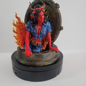 May include: A red-skinned figure with horns and a blue shirt emerges from a hatch, surrounded by flames. The figure has a red headband and is posed in front of a circular, metallic hatch. The base is black.