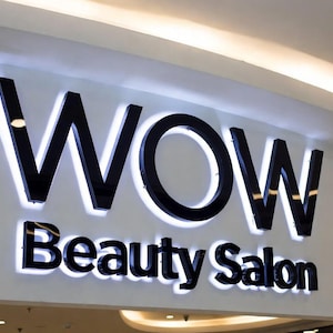 May include: A wall-mounted sign for WOW Beauty Salon featuring large, black, three-dimensional letters with a glossy finish. The letters are backlit with a bright white light, creating a glowing halo effect against the light-colored wall surface.
