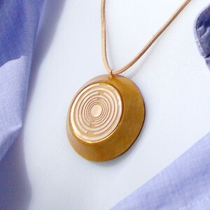 Peut inclure: Un collier pendentif en bois avec un motif circulaire. Le pendentif est de couleur marron clair et est suspendu à un cordon fin de couleur beige. Le collier est présenté sur une chemise boutonnée bleue.