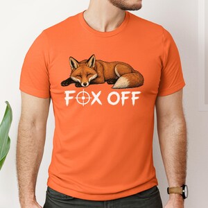 Può includere: Una t-shirt a maniche corte color arancione brillante con la grafica di una volpe marrone a riposo sopra la scritta bianca "FOX OFF", in cui la lettera "O" è sostituita da un simbolo di mirino circolare bianco al centro del petto.