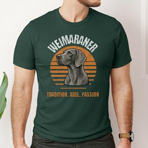 Könnte beinhalten: Ein dunkelgrünes Kurzarm-T-Shirt mit einer Grafik eines Weimaraner-Hundekopfes im Profil. Hinter dem Hund befindet sich ein orangefarbener Sonnenuntergangskreis. Der Text lautet WEIMARANER oben und TRADITION. ADEL. PASSION unten in Orange.