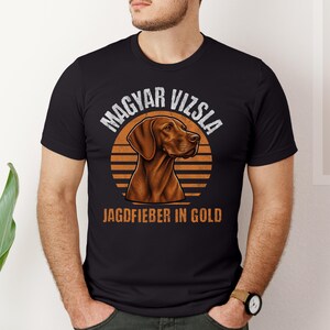 Op de afbeelding: Een zwart T-shirt met korte mouwen en een afbeelding van een bruine Vizsla-hondenkop, gecentreerd boven een oranje zonsondergangcirkel. De tekst MAGYAR VIZSLA staat in een boog boven de hond en JAGDFIEBER IN GOLD is in het oranje afgedrukt.