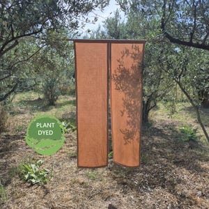 Op de afbeelding: Twee rechthoekige panelen van lichtoranje stof met een zichtbare geweven textuur. De panelen zijn voorzien van een donkerdere oranje rand langs de zijkanten. Een groen rond embleem met de tekst PLANT DYED staat linksonder op de stof.